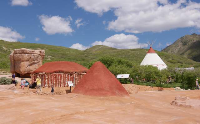 A Native American hogan and other village items with a teepee in the background surrounded by green hills and blue sky