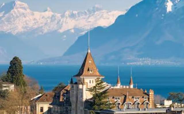 Swiss style buildings with a lake in the background that has mountains and snow covered peaks on the other side
