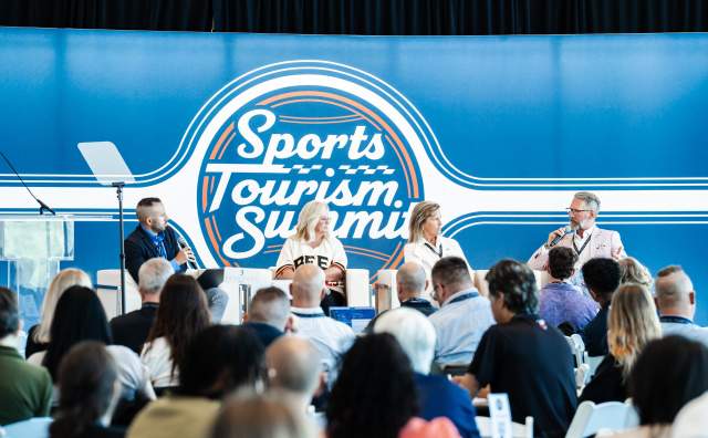 Audience seated at tables looking at stage with 4 people seated in white chairs with a Sports Tourism Summit background