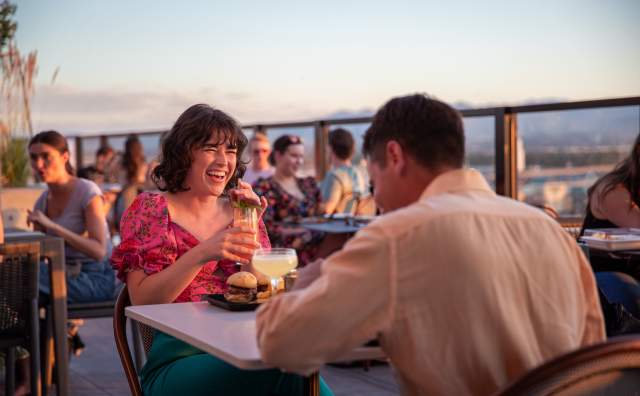 A man and a woman having dinner at a table. The woman is smiling and laughing. The background is a sunset shot over Salt Lake
