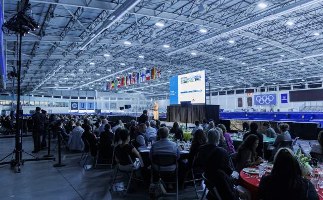 Speaker on stage at the Utah Olympic Oval, people sitting at round tables in the foreground, UT Hockey Club and Olympic logos in the background