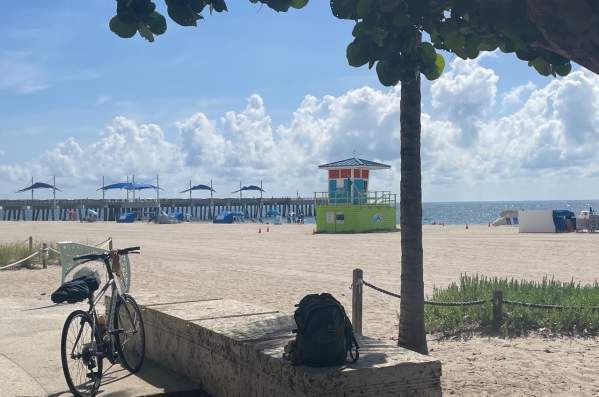 Bicycle standing at the edge of the beach with the Pompano Beach Pier and a lifeguard tower in the background.