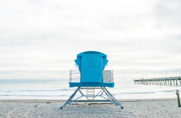 Blue lifeguard tower positioned on a sandy beach in Dania Beach, with palm trees nearby, gentle ocean waves in the background, and a long fishing pier extending into the water.
