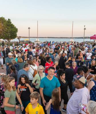 A crowd of hundreds of people dance on the rooftop of Monona Terrace with Lake Monona in the background