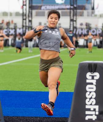 A woman with a gray shirt and green shorts leaps across the finish line at the 2021 CrossFit Games