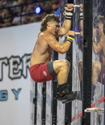 A man with a mullet and red shorts climbs up a plexiglass pegboard at the 2021 CrossFit Games