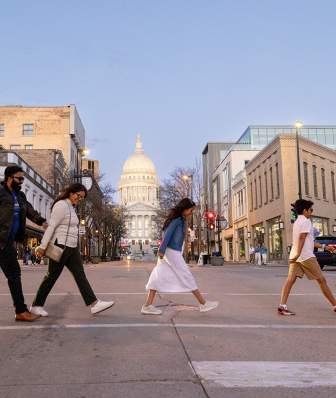 A family of four visiting Madison, WI walks across the street with the Wisconsin State Capitol and art museum in the background