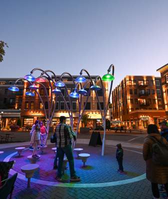 Colorful light fixtures illuminate a lively plaza at dusk, with people interacting and enjoying the vibrant atmosphere near shops and restaurants.