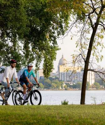 Two bikers ride along the Monona Lake Loop on a sunny day with the Capitol visible in the background across the lake.