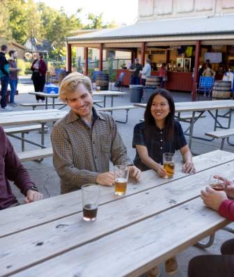 Four friends sit at a wooden picnic table outdoors, enjoying drinks in a lively setting with people and a bar in the background.