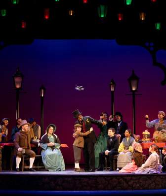 A vibrant stage scene featuring a diverse cast in period costumes, gathered around tables with lanterns above, set against a colorful backdrop.