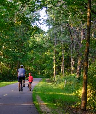 An adult and a child bike along a paved, wooded trail