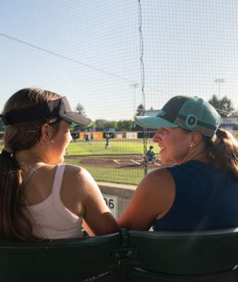 Two people sit in stadium seats, talking while watching a softball game on a sunlit field behind a protective net.