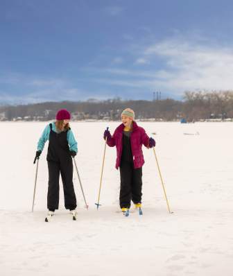 Two girls laughing and cross country skiing next to each other across a frozen, snow covered lake.