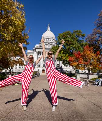 Two women wearing red and white striped overalls posing in front of the Wisconsin State Capitol building.