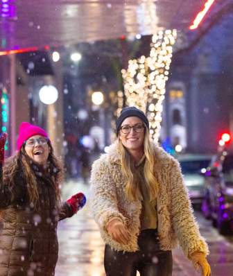 Two women walking and throwing snowballs in downtown Madison as the trees are lit up with holiday lights.