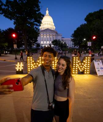 Two people take a selfie in front of the light up MNM sign in front of the Capitol building