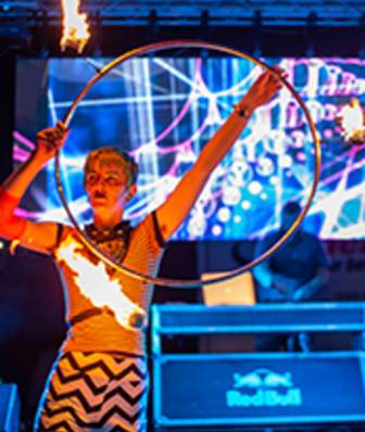 A white woman holds a hoop that has flaming balls attached arounf the sides. The woman is standing on a dark stage with neon colors behind her