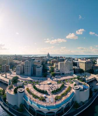 Aerial view of a vibrant city skyline along a river, featuring modern buildings, green spaces, and a clear blue sky. Sunlight reflects on the water.