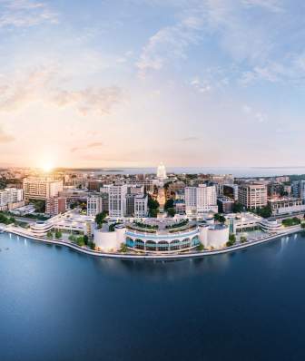 A panoramic view of a vibrant city skyline along a waterfront at sunrise, showcasing modern buildings and lush greenery.