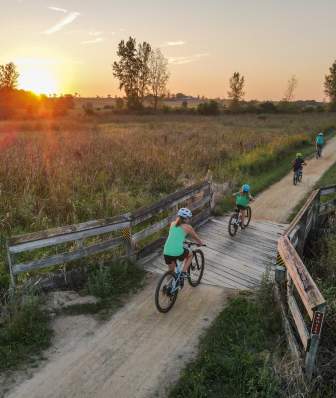 Four bikers cross a wooden bridge surrounded by green pastures. The sun is setting in the distance.