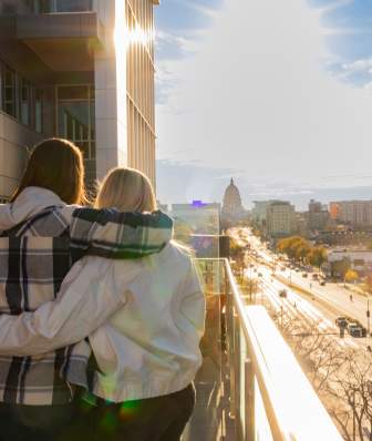 Two women stand on a balcony, overlooking a cityscape bathed in sunlight. The view includes buildings and a distant dome, with a busy street below.
