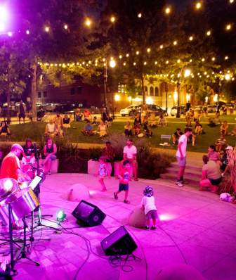 A band plays on an outdoor stage at a downtown park surrounded by lights