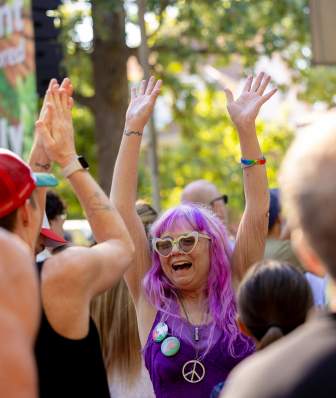 A person with long purple hair and light-colored sunglasses raises both arms in a crowd at an outdoor festival, surrounded by other attendees with hands raised near a stage set among trees.