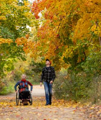 Two people travel along a wide tree-lined path in autumn, one using a wheelchair and one walking beside them, surrounded by vibrant yellow, orange and green foliage with fallen leaves covering the trail.