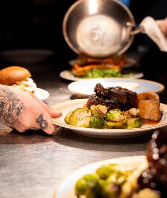 A chef slides food across the counter for a server to take and give to guests. the plate of food has a steak and Brussels sprouts covered in sauce.