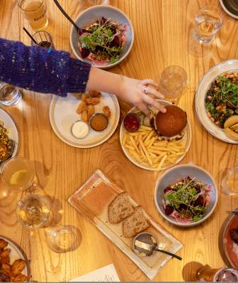 A wooden table filled with various dishes, including salads, fries, and a burger. Hands reach for food, with drinks in glasses nearby.