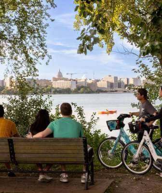 Four people sit on a bench next to two people on Bcycle e-bikes overlooking Lake Monona with the isthmus and Capitol Building in the background.