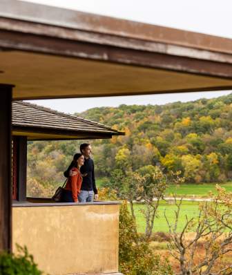 Two people stand on a covered balcony overlooking green fields and tree‑covered hills with fall foliage.