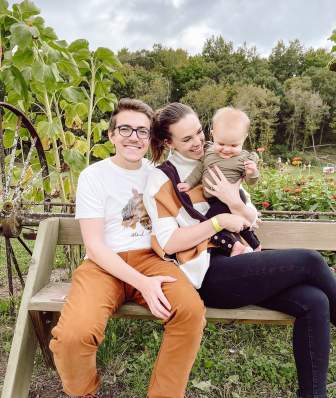 A white man, woman and baby sit on a bench outside of a corn field at Enchanted Valley Acres