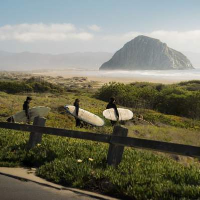 Three surfers with surf boards walking to the ocean with Morro Rock in the background