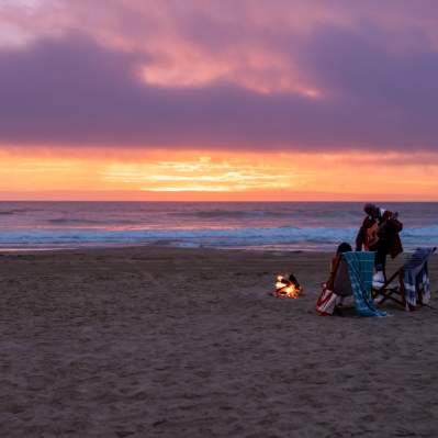 Couple on chairs at beach bonfire at sunset next to a car with ocean in background