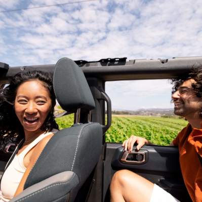 Two people smiling with open top car with wind in hair and fields in background