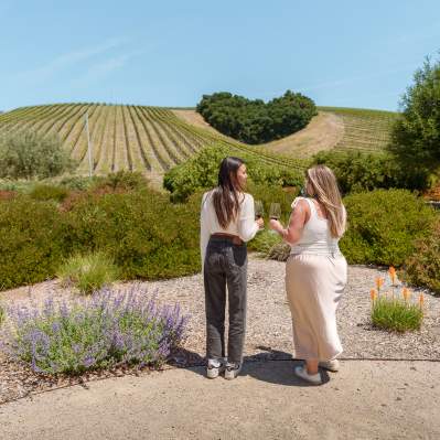 Two women tasting wine standing in a garden with hills filled with vines in the the background