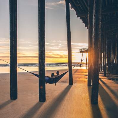 A couple relaxes in a hammock strung between pier pylons on a SLO CAL beach at sunset.