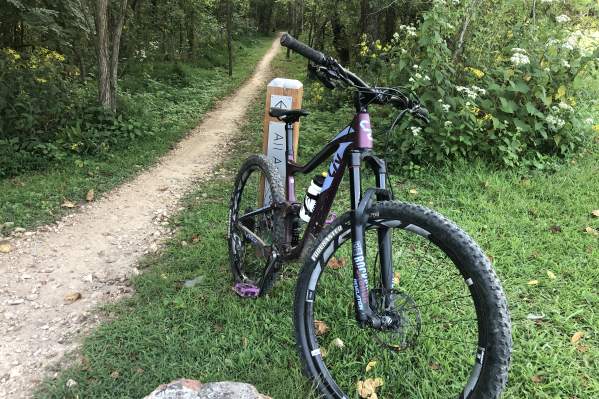 Purple mountain bike leaned against a trail marker that reads "All American"