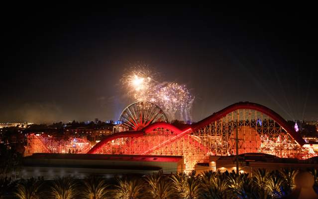 Disney fireworks lighting up the night sky over a brightly illuminated roller coaster and Ferris wheel at a theme park in Anaheim, California.