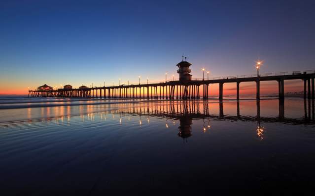 Huntington Beach pier at sunset with lights reflecting on wet sand, extending into calm ocean waters under a gradient sky of deep blue and warm orange hues.