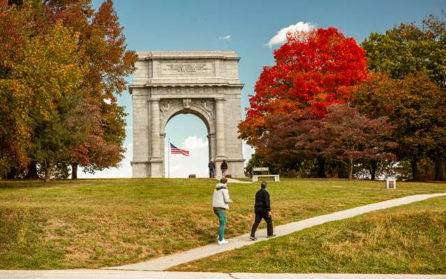 An older male and female walk casually up a hill in front of the monument arch at the Valley Forge National Historic Park surrounded by trees with fall foliage.
