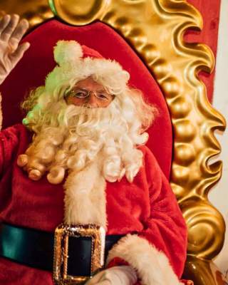 A jolly looking Santa waves as he sits on a plush throne in his grotto
