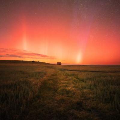 A barn sits in a field beneath a red night sky full of stars.