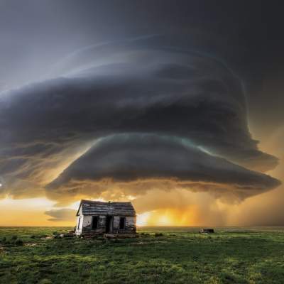 A towering supercell storm glows at sunset over eastern New Mexico grasslands near an abandoned farmhouse.