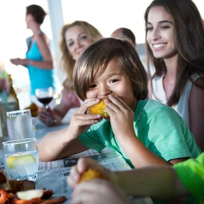 A child looks into the camera and bites into a fresh piece of corn