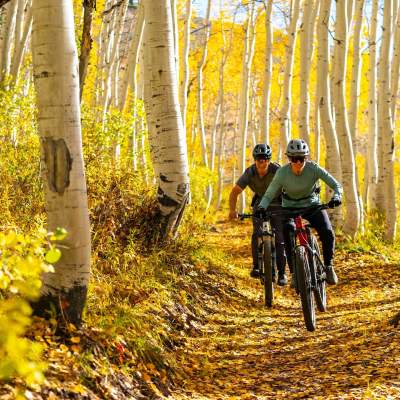 Two riders smile as they ride their bike through fall leaves on a trail near Park City, UT