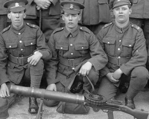 A Welsh regiment of soldiers kneel behind a Lewis Gun. A row of soldiers stand behind them with only their feet visible. The soldiers are in First World War army uniforms and hats.