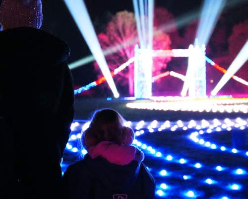 A mother and small child stand with their backs to the camera looking at a small, lit up London Bridge. There are roaming spotlights & glowing fairylights on the floor in all different colours. The trees off in the distance are also lit up in blue and pink.
